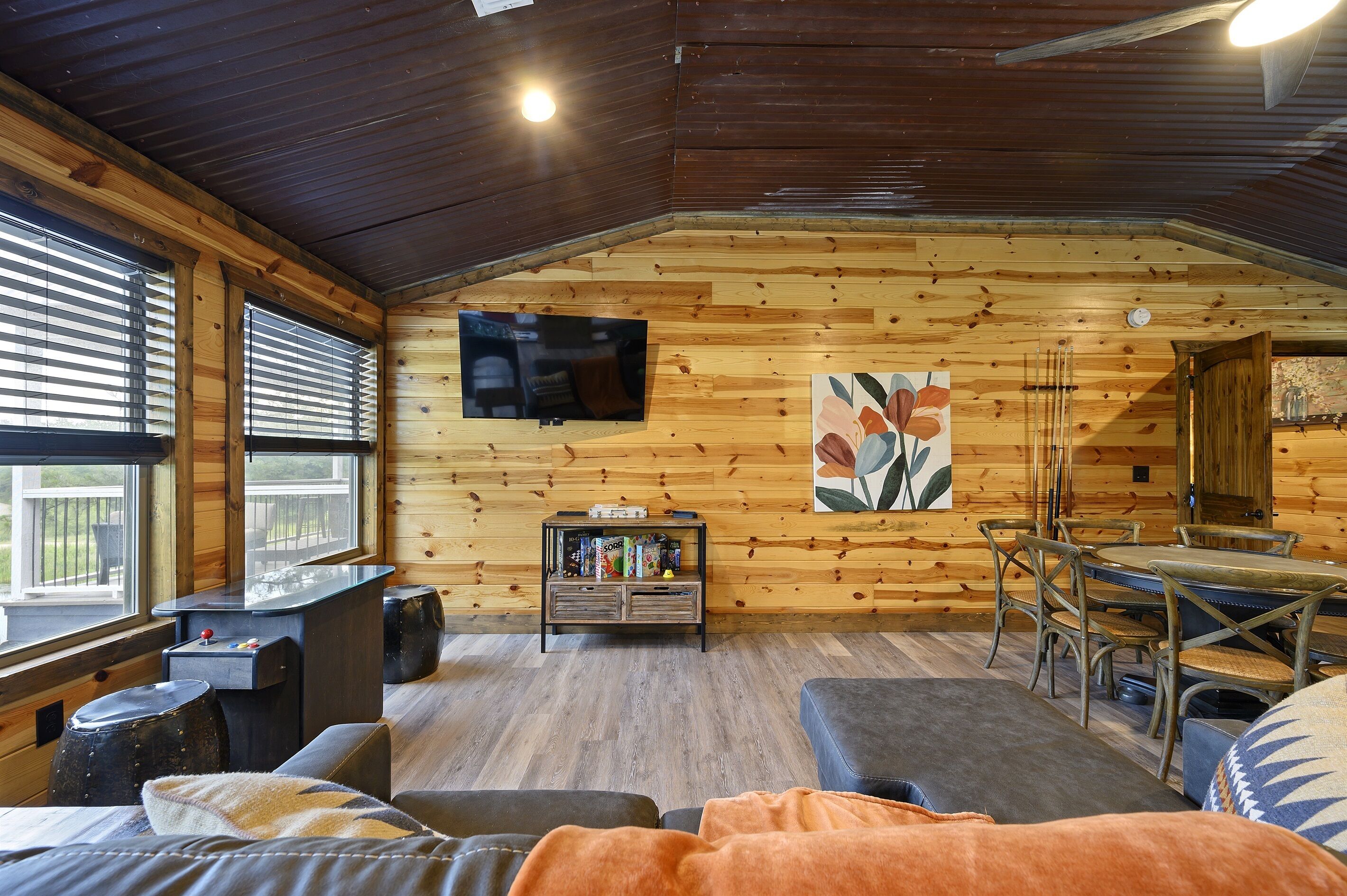 Interior acogedor de una cabaña con detalles de madera, un televisor montado en la pared, una zona de comedor y un rincón de descanso con cojines.