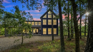 A modern two-story house with large windows and a symmetrical design, surrounded by a forested area.