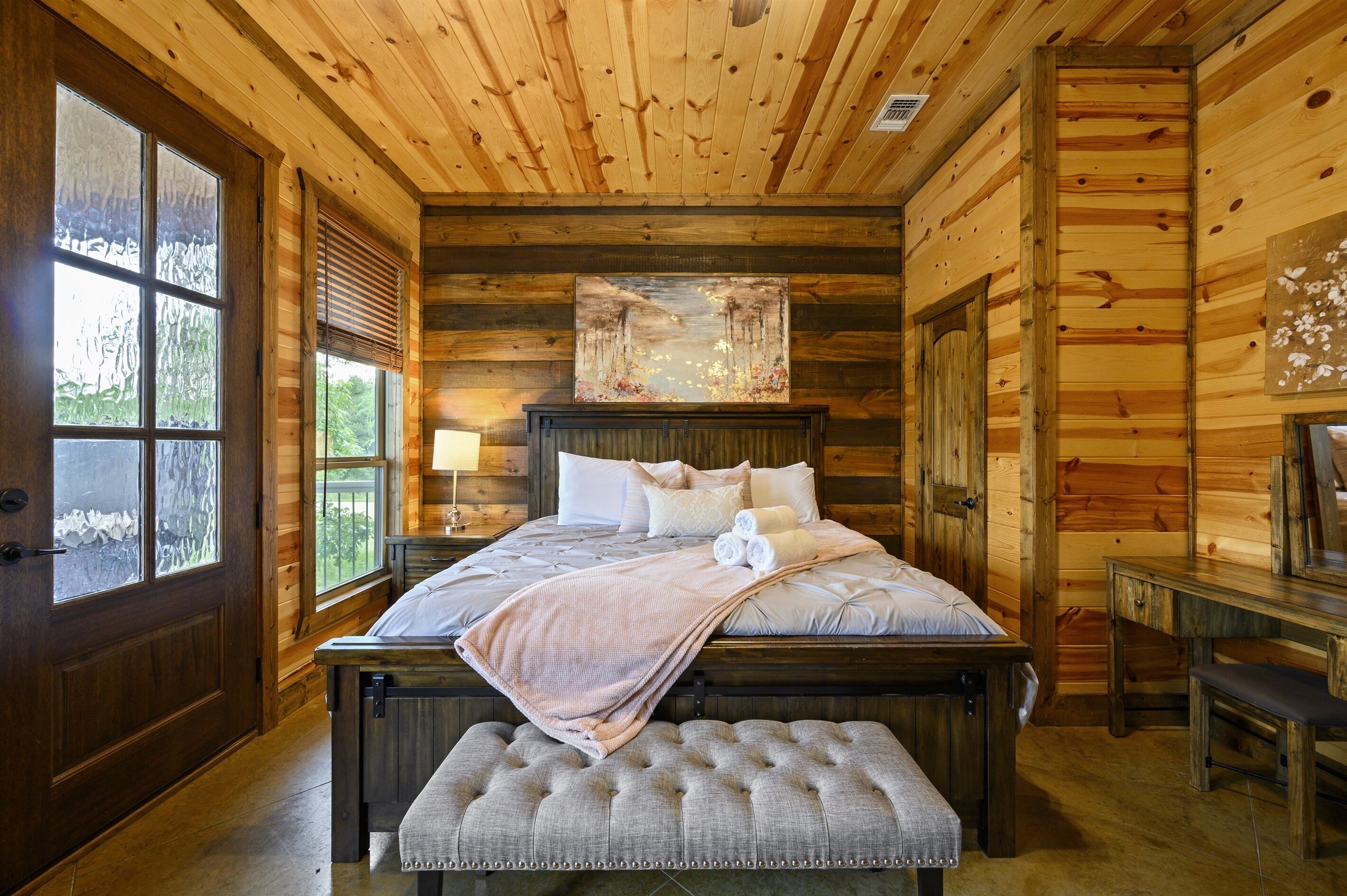 A wooden cabin bedroom with a large bed, a bench, and a painting on the wall.