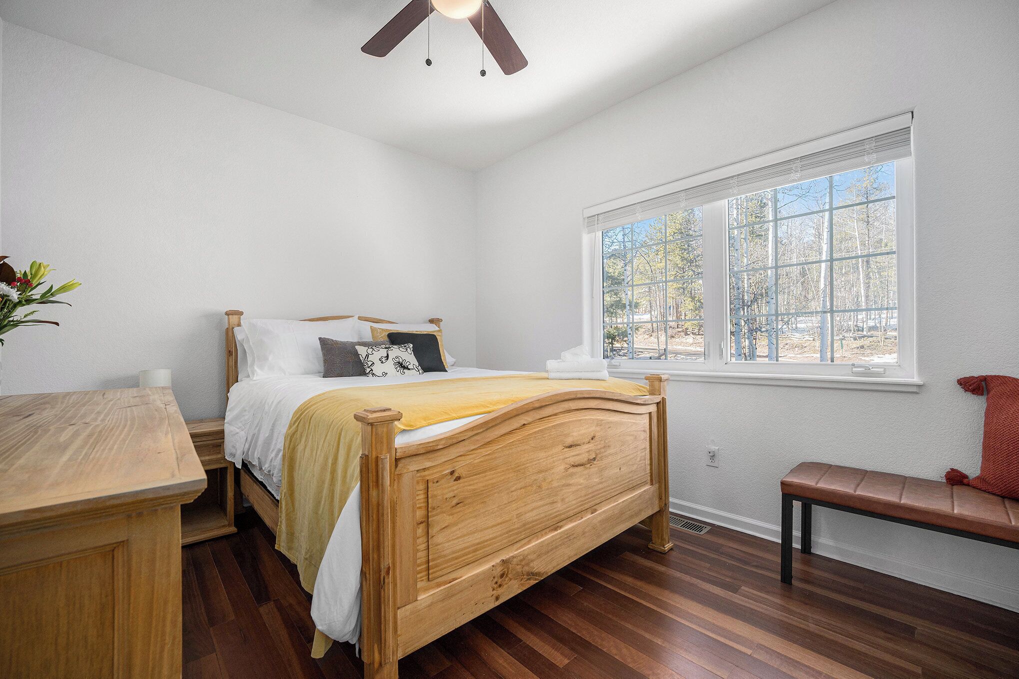 Een slaapkamer met een houten bed, een houten bureau, een bankje en een raam met uitzicht op bomen.