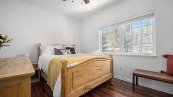 Een slaapkamer met een houten bed, een houten bureau, een bankje en een raam met uitzicht op bomen.