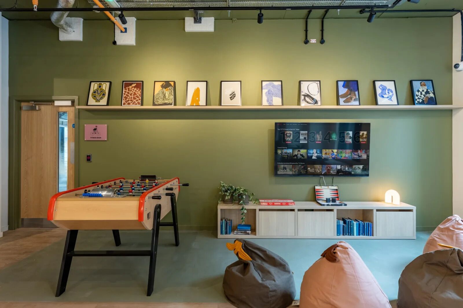 A room with a foosball table, a shelf with framed artwork, a TV displaying a movie, and a bookshelf.