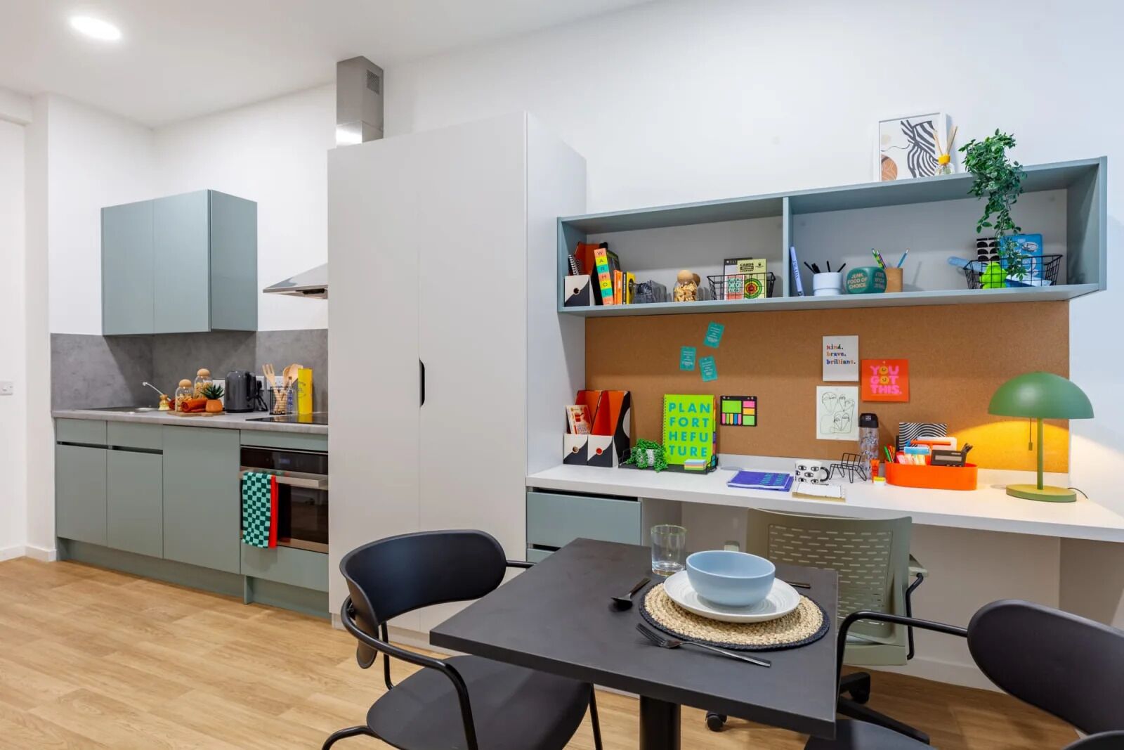 Una cocina moderna con armarios grises, un comedor con mesa y sillas, y una zona de trabajo con estanterías y un tablón de corcho.