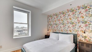 Une chambre à coucher avec un grand lit, une fenêtre, un papier peint à motifs floraux et une table de chevet avec une lampe.
