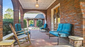 A covered patio with brick walls, two rocking chairs, and two blue cushioned chairs. - Spacious Historic Manor with Chef's Kitchen • Horse-Friendly (Southern Pines)