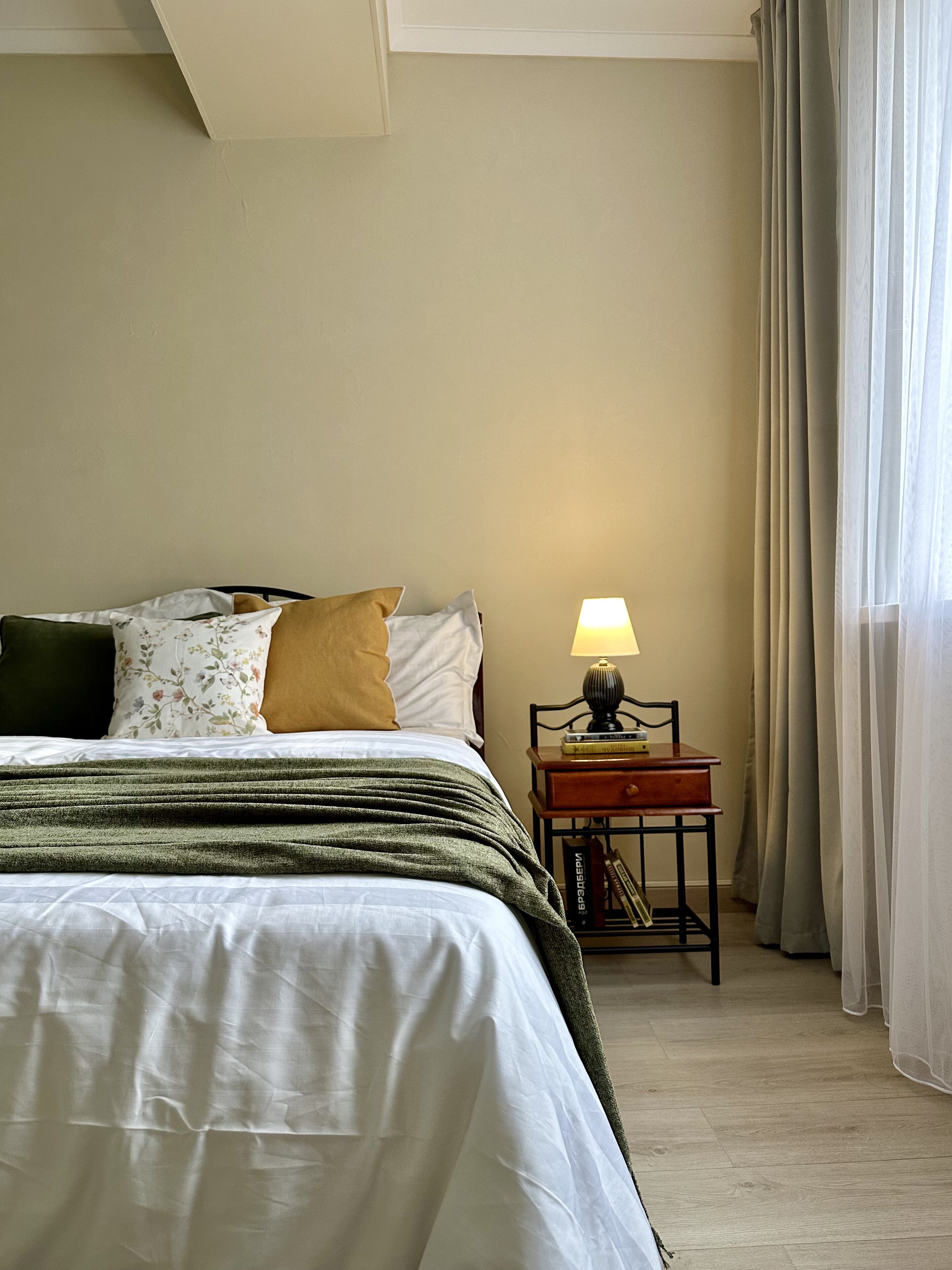 A neatly made bed with a green blanket, a bedside lamp, and a small wooden nightstand with books.