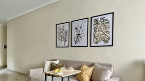 Un salon avec un canapé beige, une petite table en bois sur laquelle se trouvent un livre et un bol de fruits, et trois reproductions botaniques encadrées accrochées au mur.