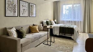Une chambre d’hôtel moderne avec un canapé, une petite table avec une lampe, un lit recouvert d’une couverture verte et trois reproductions encadrées de motifs botaniques accrochées au mur.