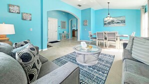 A living room with a blue wall, a dining area with a table and chairs, and a kitchen in the background. - Highgate Haven - Bright pool home with games room in gated community (Davenport)