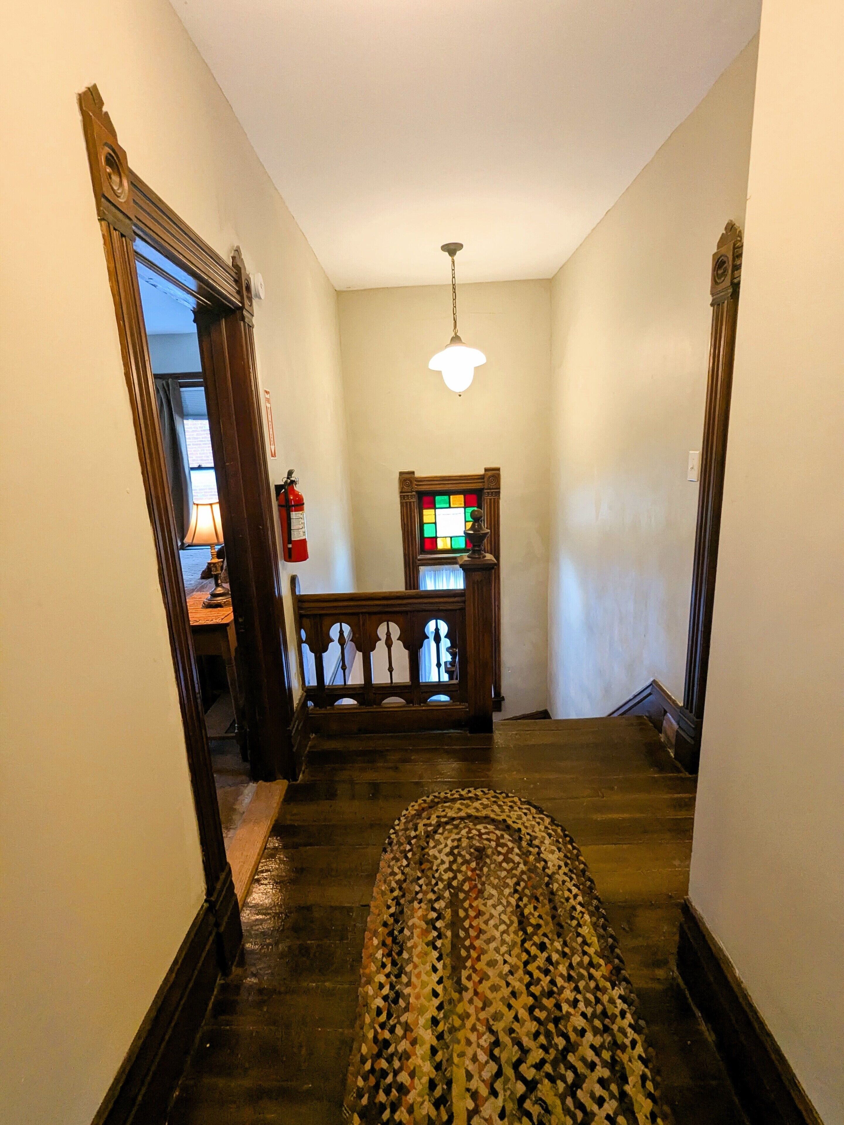 A hallway with a patterned rug, wooden staircase, a small table with a lamp, and a wall-mounted clock.