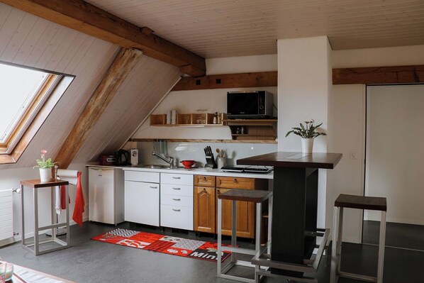 Eine kompakte Küche mit weißen Schränken, einer Mikrowelle und einem kleinen Kühlschrank. Ein Holzbalken-Boden und ein Fenster sorgen für natürliches Licht.