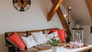 Un salón acogedor con techo de madera, un sofá de cuero marrón con cojines rojos y blancos, una mesa de centro de madera con un jarrón blanco y vasos, y un reloj de pared.