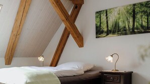 Un dormitorio con techo inclinado, una cama con manta verde, una mesita de noche con lámpara y un cuadro de arte con tema de bosque.