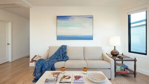 Ein Wohnzimmer mit einem beigen Sofa, einem Couchtisch mit Zeitschriften, einer blauen Decke und Meerblick durch ein Fenster.