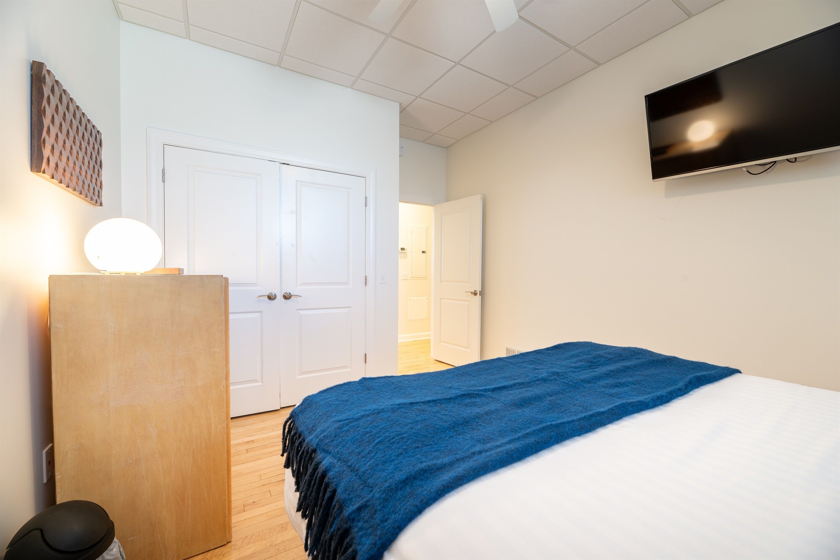 A bedroom with a bed, a TV mounted on the wall, a wooden dresser, and a door leading to another room.