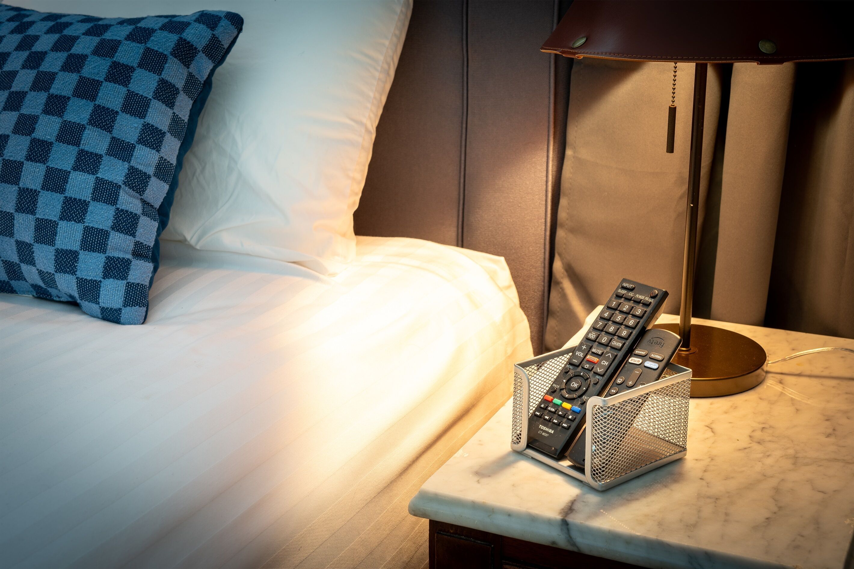 Ein ordentlich gemachtes Bett mit einem karierten Kopfkissen, einer Fernbedienungshalterung und einer Lampe auf einem Nachttisch.