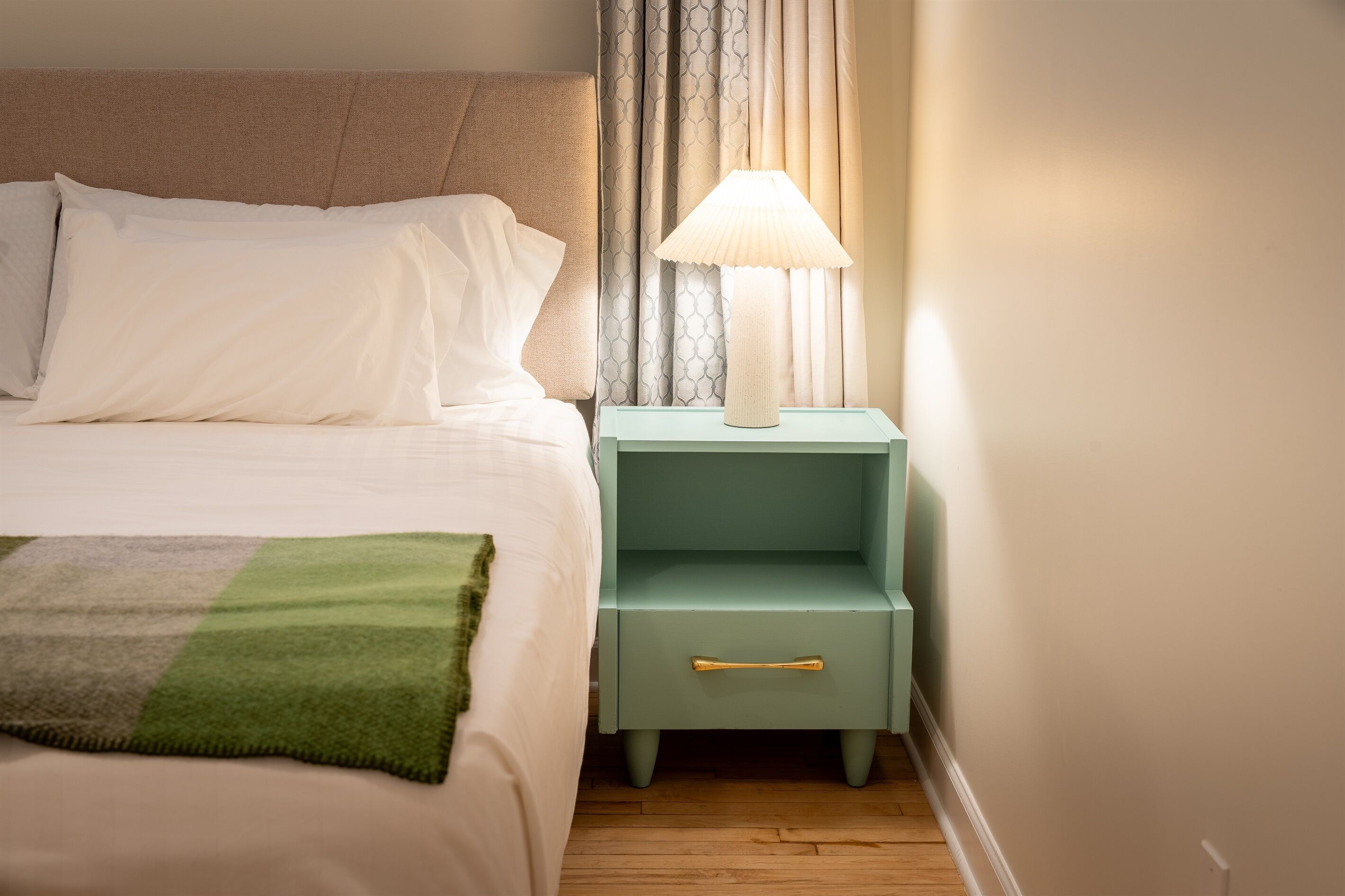 A neatly made bed with white linens, a green throw blanket, and a nightstand with a lamp.