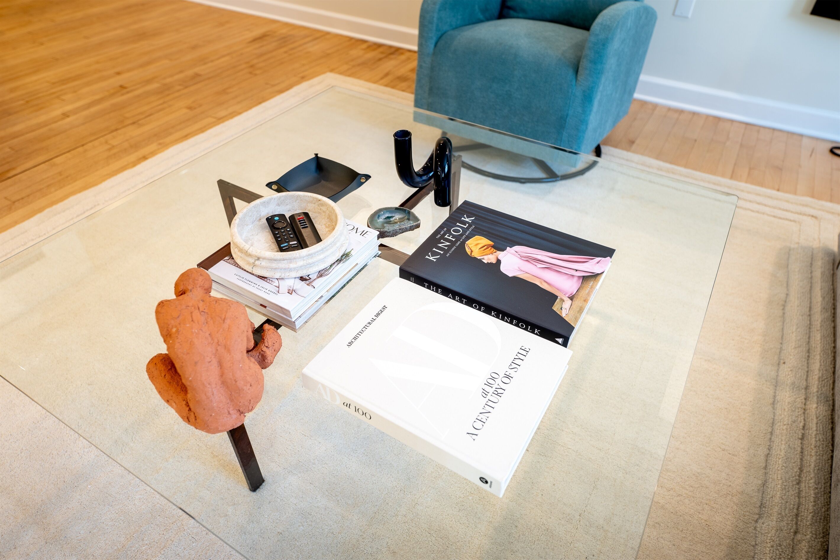 A living room with a glass coffee table, a blue armchair, and a book titled "The Art of Kinfolk".