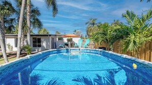 Una piscina amb una terrassa de fusta, envoltada de palmeres i una casa al fons.