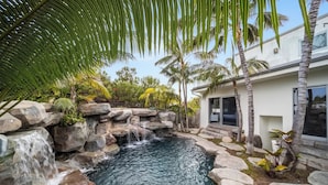 A pool with a waterfall, surrounded by rocks and palm trees, with a modern house in the background. - Encinitas Shores - Beachfront, Ocean View & Patio (Encinitas)