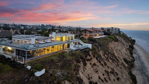 A modern coastal house with a glass facade, a spacious outdoor area with seating, and a view of the beach and ocean. - Encinitas Shores - Beachfront, Ocean View & Patio (Encinitas)