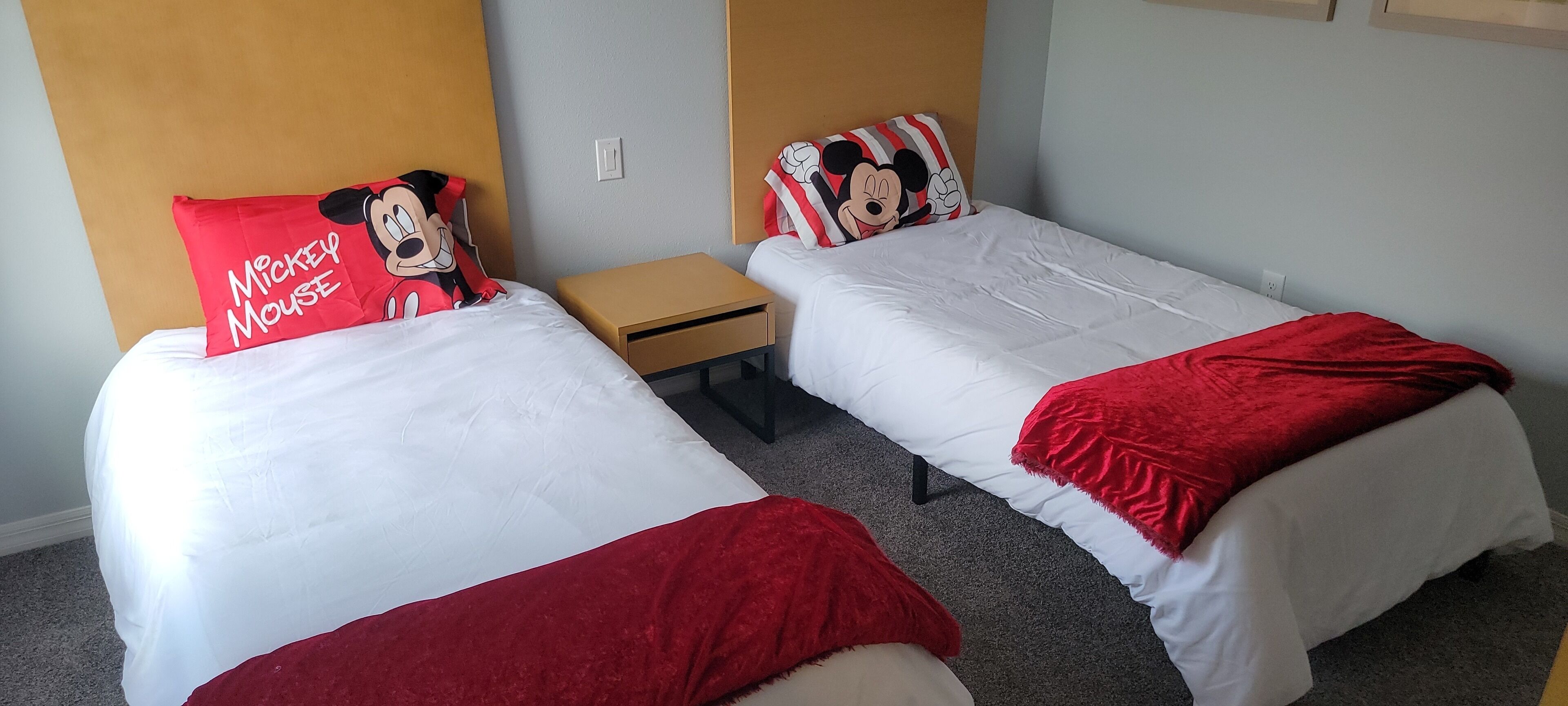Two single beds with Mickey Mouse pillows, a small wooden nightstand, and a red throw blanket.
