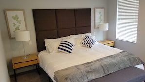 Une chambre à coucher avec un grand lit, deux tables de chevet, une commode, une fenêtre avec des stores et des lampes fixées au mur.
