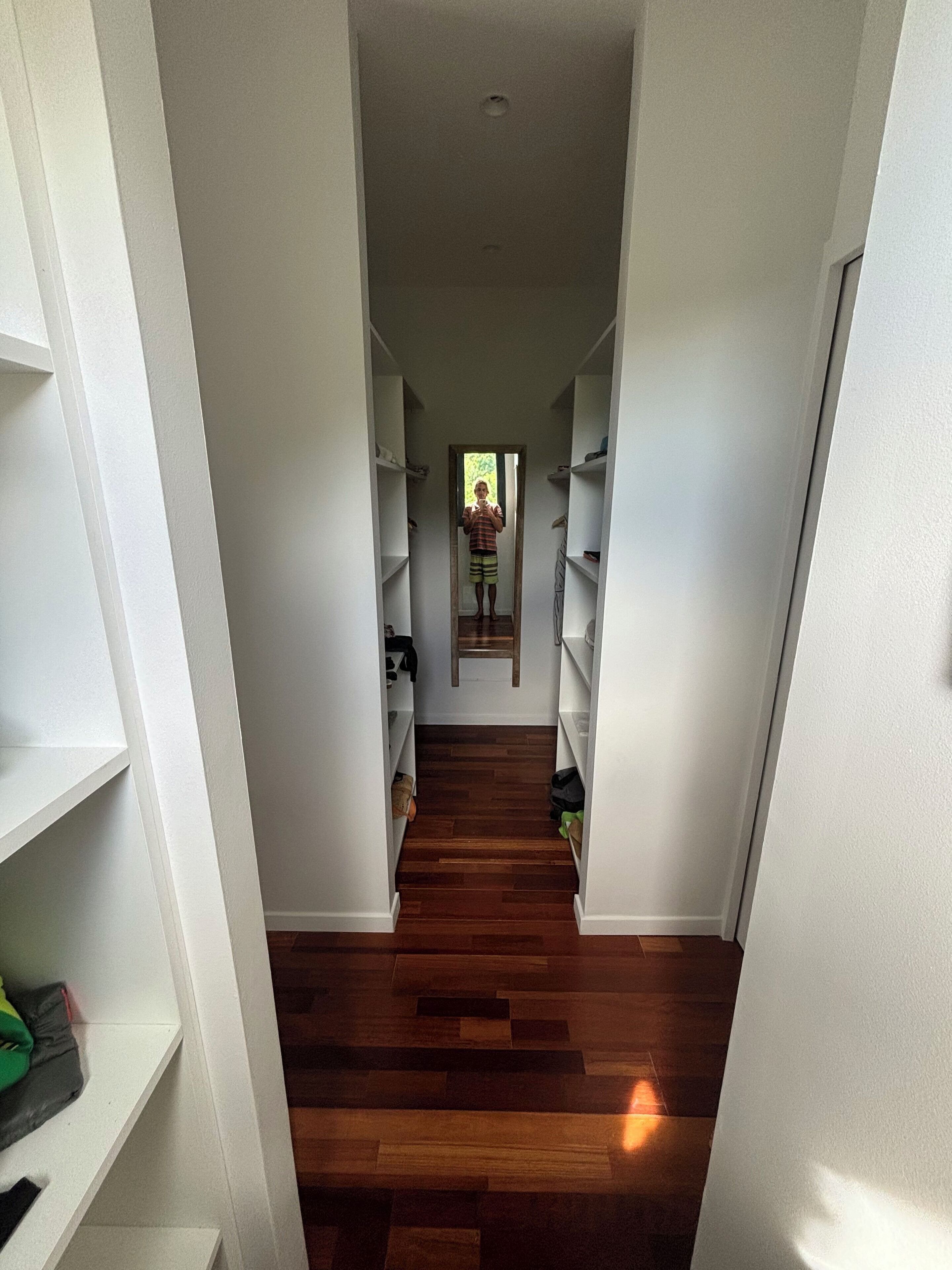 Un couloir étroit, doté d’un plancher en bois et de murs blancs, mène à une porte au fond. Des étagères sont disposées le long des deux murs du couloir.