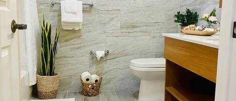 A bathroom with a marble wall, a toilet, a sink with a wooden vanity, a potted plant, a wicker basket with toilet paper, and a beige rug.