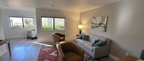 A living room with a sofa, armchair, and a rug. There are windows and a painting on the wall.