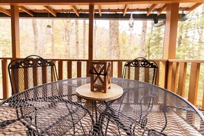 Une table ronde en métal, dont le plateau est constitué d’un motif tissé, entourée de quatre chaises dont les dossiers présentent des motifs complexes, le tout installé sur une terrasse en bois offrant une vue sur les arbres.