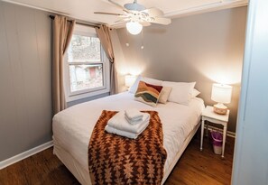 Un lit bien fait, recouvert d’une couverture marron, un ventilateur de plafond, une fenêtre avec des rideaux et une lampe de chevet.