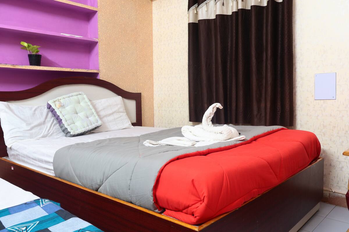 Una cama bien hecha con un edredón rojo, una almohada blanca y una almohada decorativa. Hay una pequeña planta en maceta sobre una estantería morada.