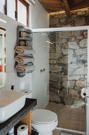 A bathroom with a stone wall, a glass shower enclosure, a white toilet, and a towel rack with neatly rolled towels. - Villa Mosaico (Real de Catorce)