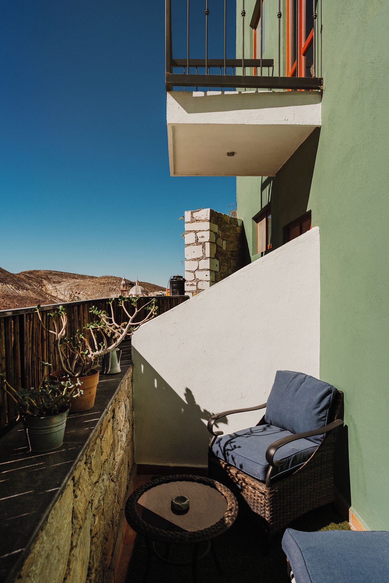 A balcony with a wicker chair and a small table, a potted plant, and a view of the hills.