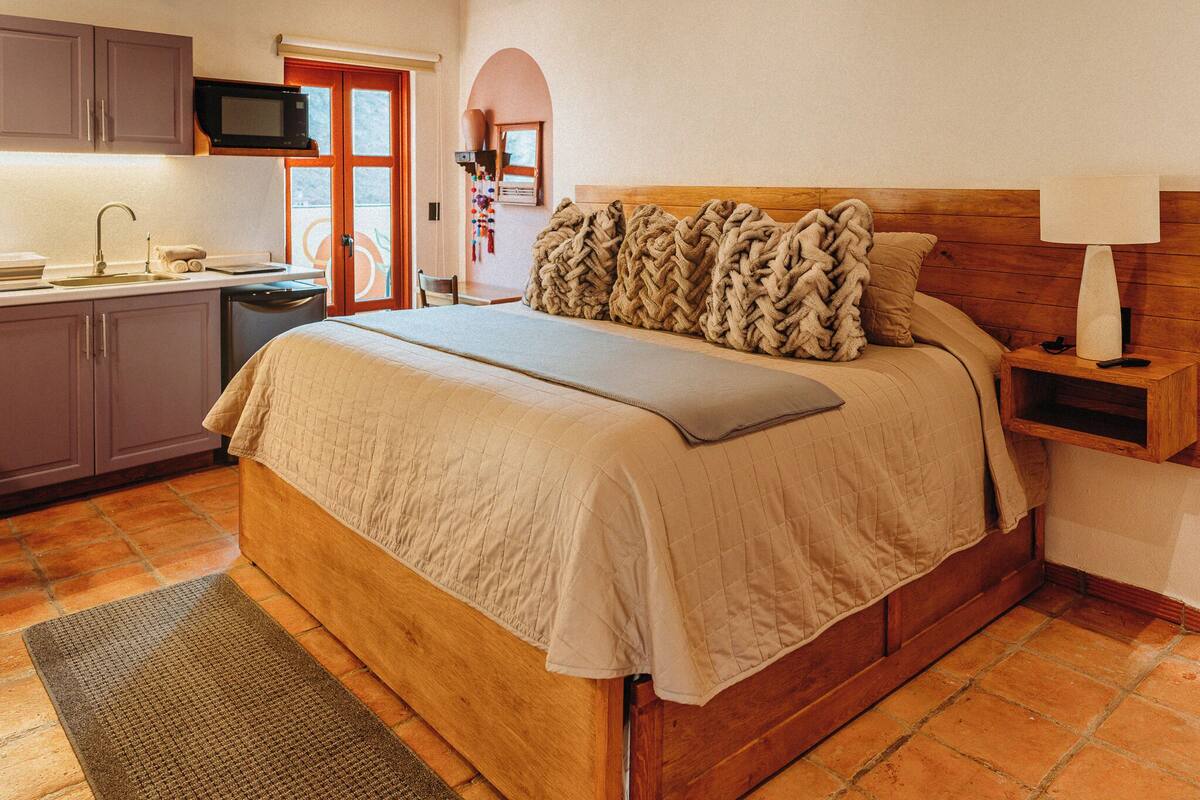 A bedroom with a wooden bed, a bedside table with a lamp, a small desk with a chair, and a kitchen area with a microwave.