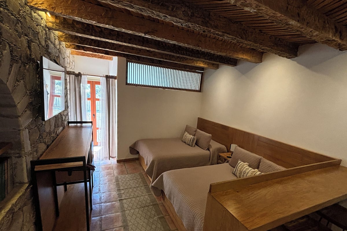 A cozy room with a stone wall, wooden ceiling beams, two beds, a wooden table, and a view of the outdoors.