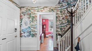 A hallway with patterned wallpaper, a wooden floor, a red wall, a clock, and a staircase with a coat rack. - Spacious 1BR on Salem Common l Steps to Downtown l Fenced in Yard + Parking (Salem)