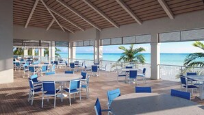 Een overdekte eethoek buiten met tafels en stoelen, met uitzicht op een strand.