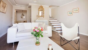 Ett modernt vardagsrum med en vit soffa, ett vitt soffbord med en vas med blommor, en vit stol, en kristallkrona och en trappa.