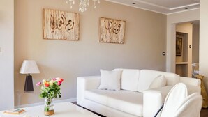 Ett modernt vardagsrum med en vit soffa, ett glasbord med en vas med blommor, en svart metallstol, trägolv och en kristallkrona.