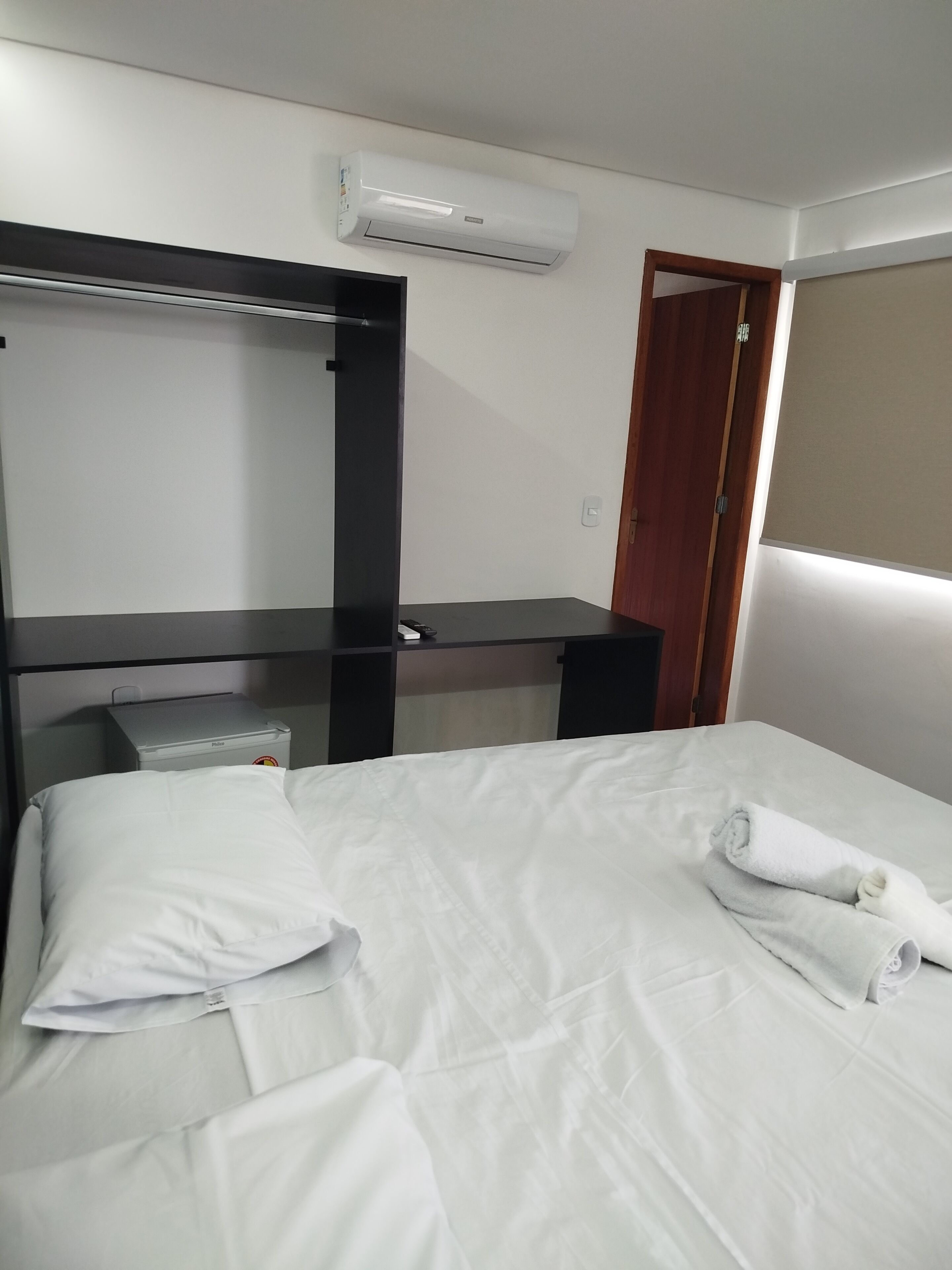 A hotel room with a bed, a desk with a chair, an air conditioner, and a door.