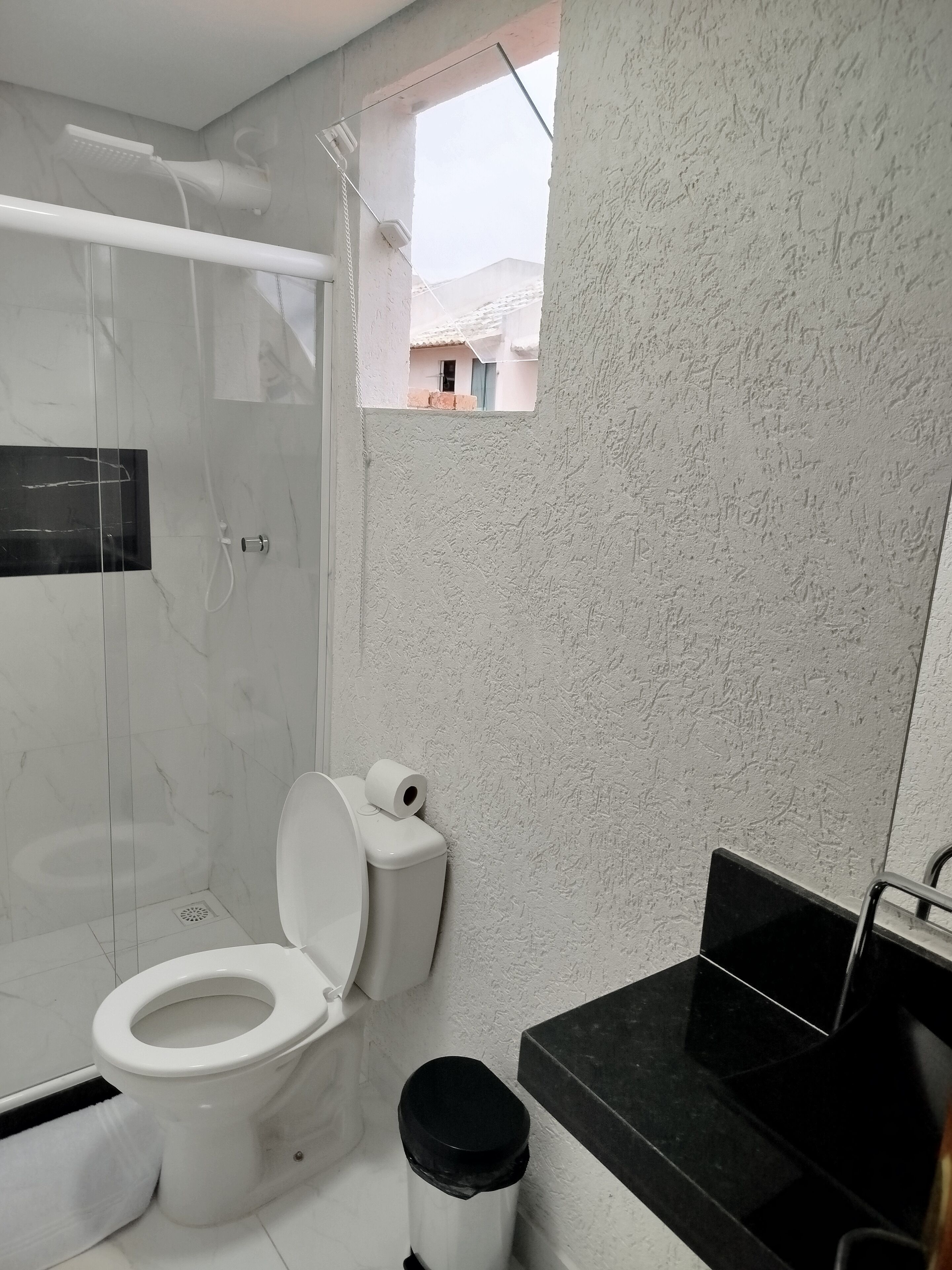Une salle de bain comprenant des toilettes, un espace douche, une petite fenêtre et un lavabo noir.
