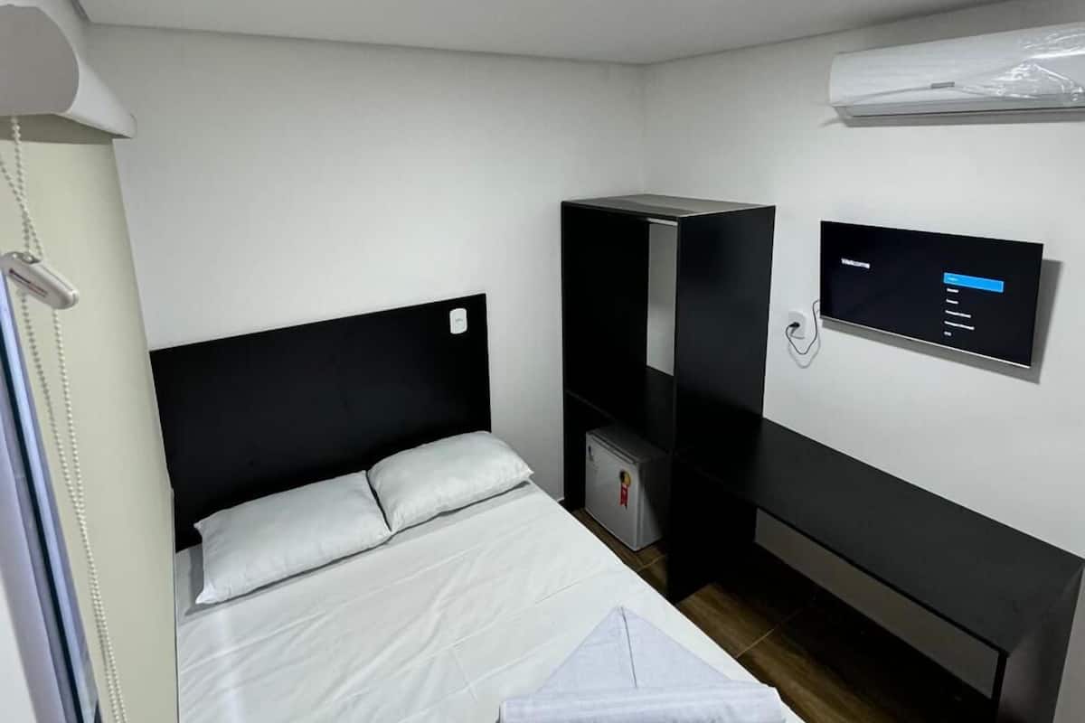 A hotel room with a bed, a desk, a small refrigerator, an air conditioning unit, and a digital display on the wall.