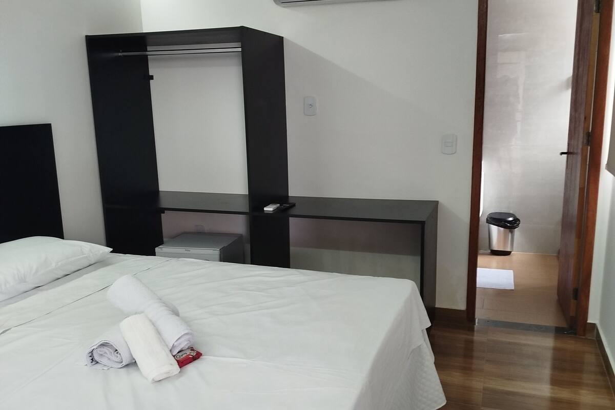 A hotel room with a bed, a desk, a mirror, an air conditioner, and a trash bin.