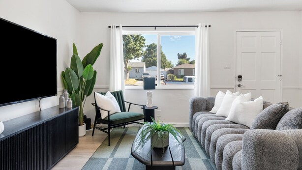 A modern living room with a flat-screen TV, a large potted plant, a sofa set, and a view of a residential area through a window. - NEW Tranquil Escape - Hot Tub, BBQ, Near LA & Disney (Covina)