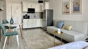 Een moderne keuken met witte kastjes, een zwarte magnetron en een grijze koelkast. Er is een eettafel met stoelen, een bank en een klein bijzettafeltje met een vaas.