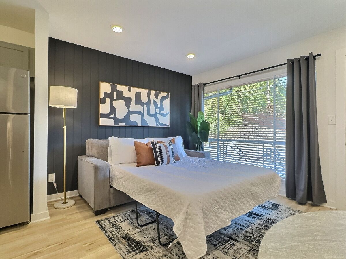 A modern bedroom with a large bed, a sofa, a floor lamp, and a view of greenery outside.