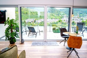 Una habitación moderna con un gran ventanal corredero que da a una terraza exterior. En el interior, hay un sofá, una mesa de comedor con sillas y una planta en maceta.