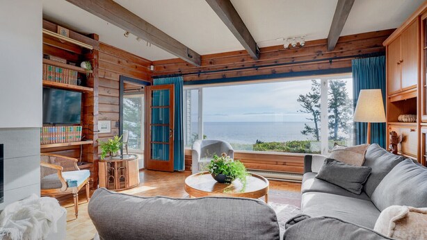 Un salón acogedor con un amplio ventanal con vistas al mar, vigas de madera en el techo, una chimenea y una estantería llena de libros.